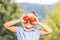 Close up shot of cute smiling child in blue dress, holding apples in front of her eyes over blurred garden background.