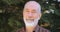 Close-up shot of face of pleasant elderly man with beard in shirt, standing in the park and smiling at the camera