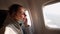 Close-up shot of happy female passenger in protective medical face mask looking in airplane window, enjoying amazing