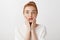 Close-up shot of impressed sympathizing redhead woman with bun hairstyle, squeezing lips while holding palms on cheeks