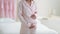 Close up shot of slim unrecognizable woman in pajamas stroking her belly, standing in bedroom at home, empty space