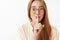 Close-up shot of tender and feminine cute redhead female in glasses with freckles showing shh sign making shush sound