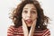 Close-up shot of touched cute feminine girlfriend with curly hair, raising eyebrows, holding palms near opened mouth