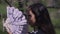 Close-up side view of gorgeous Asian young woman shaking fan looking away in sunlight in park. Headshot portrait of