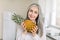 Close up of smiling elderly gray haired woman in white shirt, holding fresh pineapple while posing to camera on light