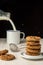 Close-up of stack of chocolate chip cookies and milk bottle serving milk in white cup, selective focus, on white wooden table, bla