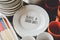 Close up stack of clean white dishes with text `HAVE, bowls, plates, mugs and colorful wooden chopsticks on metal tray