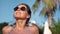 Close-up tanned face of smiling brunette woman in sunglasses enjoying sunbathing in bikini on beach