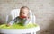 Close-up of teether toddler girl sitting on feeding chair gnawing plastic spoon. Cute infant teething and biting spoon before