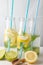 Close-up of three bottles with blue straw, water and lemon, lime and mint slices, on white table with half lemons and wooden spoon