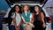 A close up of three young commonly dressed women of different nationalities sitting in an opened car trunk outside on a
