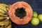 Close up to a top view of a fresh sliced orange papaya with seeds inside, a freckled bananas cluster and three green lemons over a