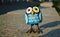 A close-up toy of a surprised blue owl with multi-colored eyes stands on a granite stone. The toy is illuminated by evening