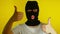 Close up of unrecognizable woman in black balaclava showing thumbs up on yellow background. Unknown female in mask