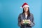 close up of an upset, disgusted Asian young woman in a Santa hat holding a green gift box, on a blue background