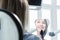 Close-up view from the back of a blonde girl sitting in a chair who looks in a tooth-shaped mirror. Reflection