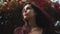 Close up view of a beautiful brunette young woman standing by the blossoming bush of red roses and looking far away