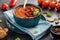 A close up view of a bowl of roasted red pepper soup surrounded by peppers, crackers and tomatoes.