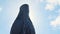 Close-up view of bronze or stone statue of priest with cross in his hand in sunny day against blue cloudy sky. Stock