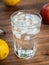 Close up view on glass full of water and melting ice cubes. Yellow lemons, red apple and glass on brown desk. Selective