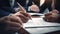 Close-up view of hands signing a document, with multiple individuals engaged in a business meeting around a table