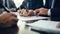 Close-up view of hands signing a document, with multiple individuals engaged in a business meeting around a table