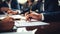 Close-up view of hands signing a document, with multiple individuals engaged in a business meeting around a table