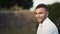 Close-up view of handsome bridegroom with brown hair and beard in white shirt looking at the camera and smiling in park