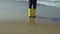 Close-up view of little girl foot in bright yellow rubber boots. Child standing on shore of beach, footprints in sand.