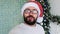 Close-up of a webcam, a portrait of a positive guy, sitting near a decorated glowing holiday tree, congratulates his
