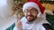 Close-up of a webcam, a portrait of a positive guy, sitting near a decorated glowing holiday tree, congratulates his