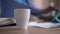 Close-up of white coffee cup on kitchen table as blurred male African American hand scrolling tablet screen at