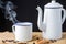 Close-up of white enameled coffee pot and cup, with smoke, coffee beans on table and black background,