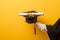 Close up of woman hand in graduation gown is holding graduation cap and certificate on yellow background