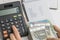 Close up of woman hand pushing button on calculator with list of budget, expense and cost on small notepad, pile of money
