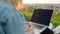 Close-up of a woman programming on a laptop sitting on a hill overlooking the city during sunset.