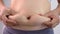 Close-up of woman`s hand pinching excessive belly fat isolated on gray background. Woman fat belly. Obesity and Overweight Concept