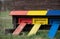 Close up of a wooden beehive. There are three openings for bees, marked in red, yellow and blue. The beehive stands in a meadow in