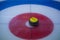 A close-up of a yellow-handled curling stone sits on ice in the center of the House