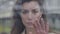 Close-up of young Caucasian woman standing behind glass and touching window with hand. Portrait of gorgeous brunette