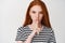 Close-up of young ginger girl with serious face showing taboo sign, hushing at you, telling a secret, white background