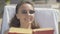 Close-up of young positive woman in sunglasses reading book at luxurious resort. Portrait of beautiful relaxed Caucasian