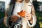 Close-up young woman hipster photographer with retro film camera holding orange fallen leaf outdoors.