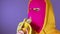Close up of young woman in pink balaclava and yellow hood eats banana on purple background. Unknown female in mask bites