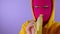 Close up of young woman in pink balaclava and yellow hood peeling banana on purple background. Unknown female in mask