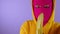 Close up of young woman in pink balaclava and yellow hood peeling banana on purple background. Unknown female in mask