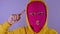 Close up of young woman in pink balaclava and yellow hood twirling finger at her temple on purple background. Surprised