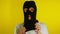 Close up young woman puts on black balaclava on yellow background. Secretive female puts on mask, looking at camera.