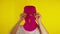 Close up young woman puts on pink balaclava on yellow background. Secretive female puts on mask, looking at camera.