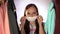 Close up of young woman putting on medical mask on her face, standing between clothes in wardrobe.